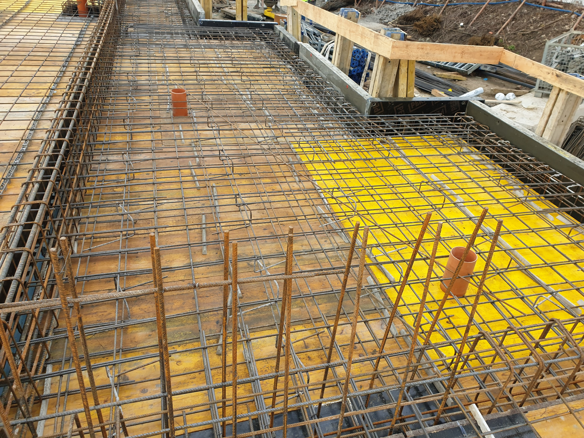 Zbrojenie płyty fundamentowej z widocznymi rurami kanalizacyjnymi, żółtą szalunkową płytą i drewnianymi elementami konstrukcyjnymi.