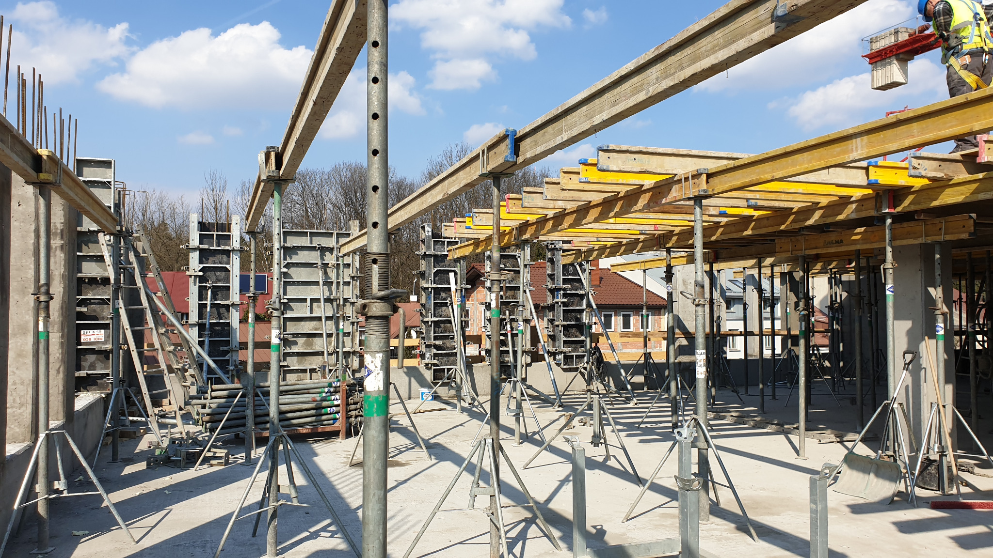Szare szalunki pionowe i poziome, wsparte na stalowych podporach z regulacją wysokości, tworzące konstrukcję na placu budowy domu jednorodzinnego w słoneczny dzień.