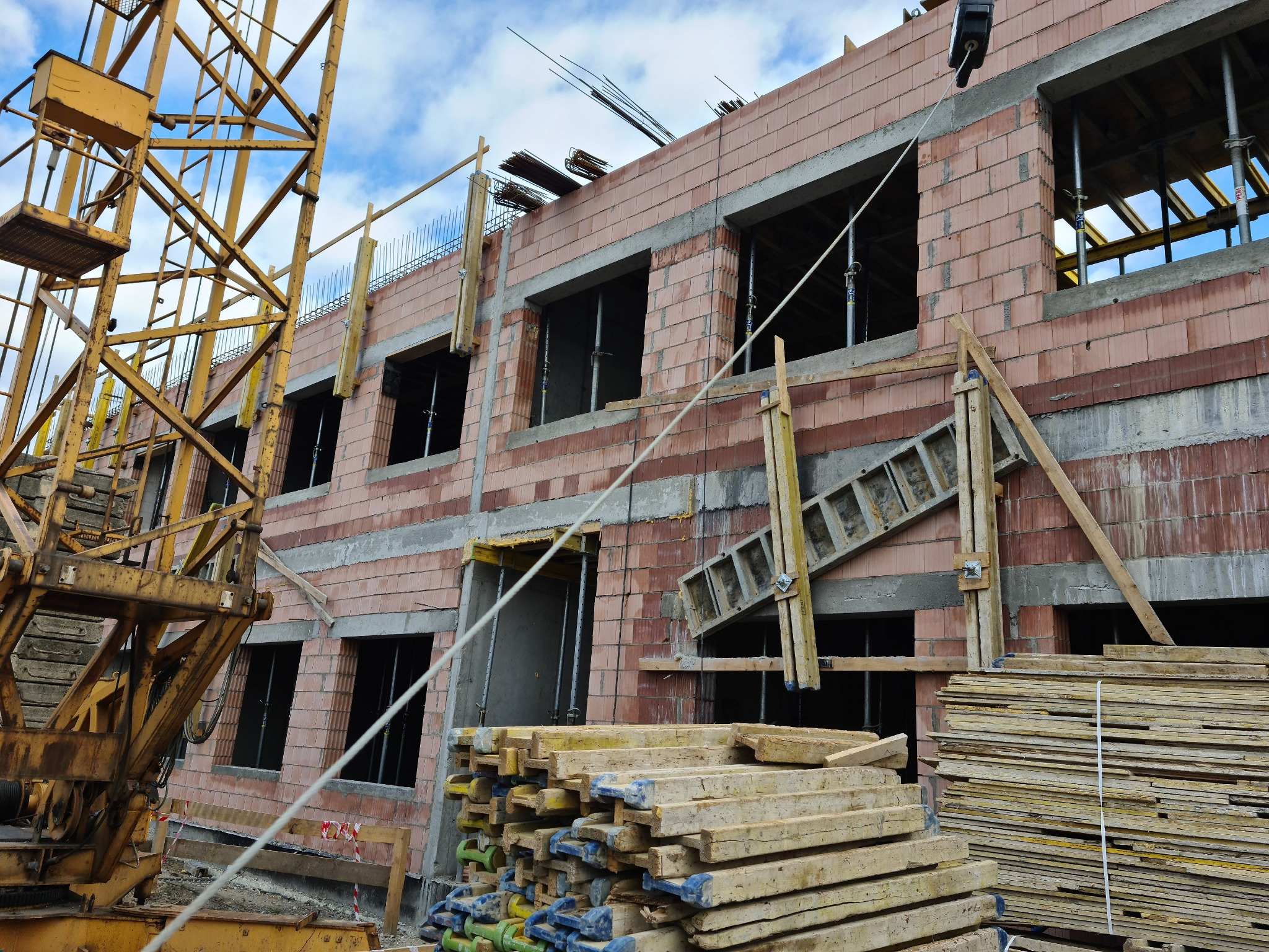 Wznoszenie dwukondygnacyjnego domu z czerwonej cegły ceramicznej, widoczne szalunki okienne, stalowe podpory, ułożone deski szalunkowe oraz żółty żuraw budowlany na tle błękitnego nieba.