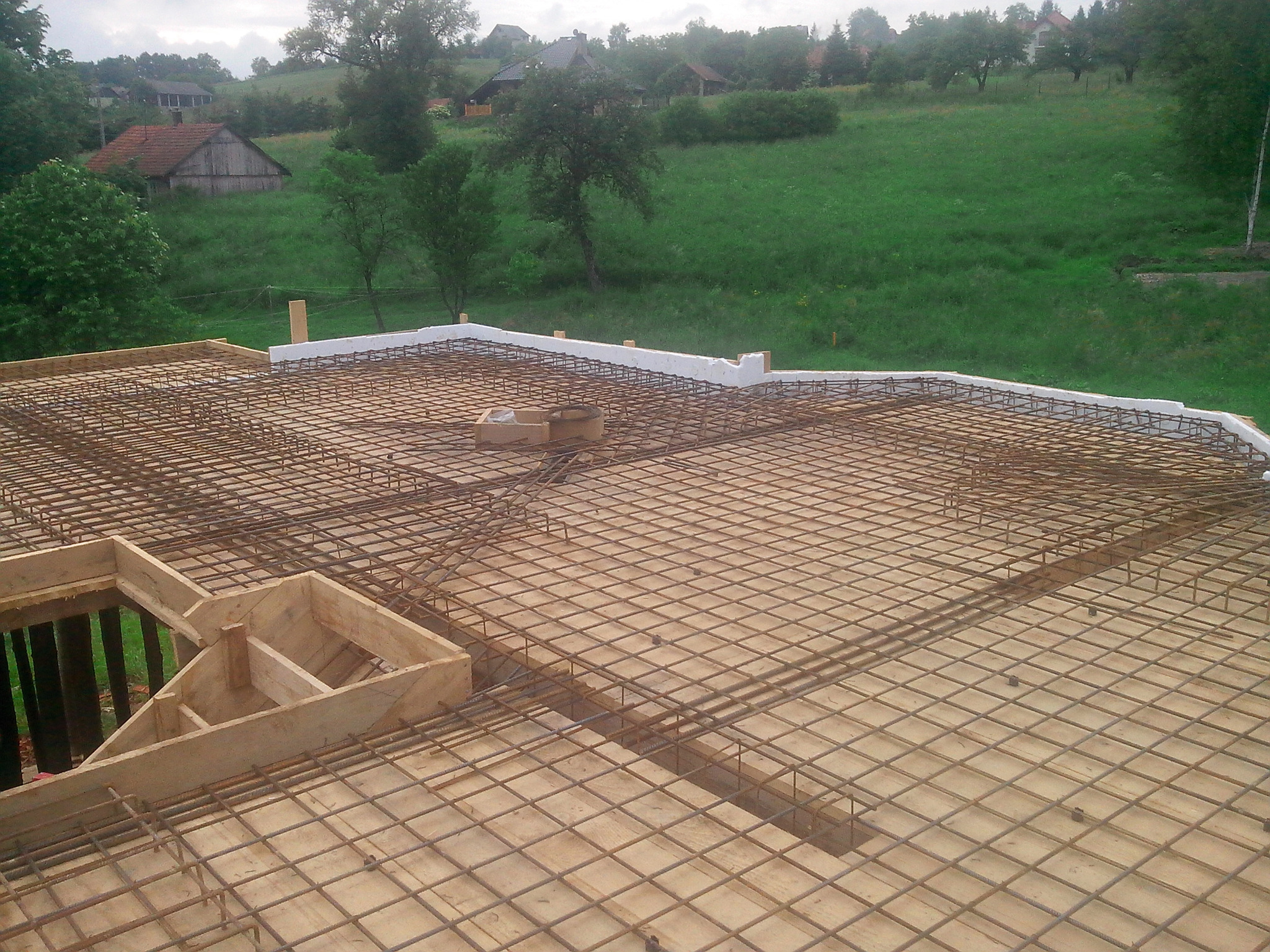 Zbrojenie płyty fundamentowej z drewnianym szalunkiem i izolacją termiczną, widok z góry na tle zielonego wzgórza z zabudowaniami.