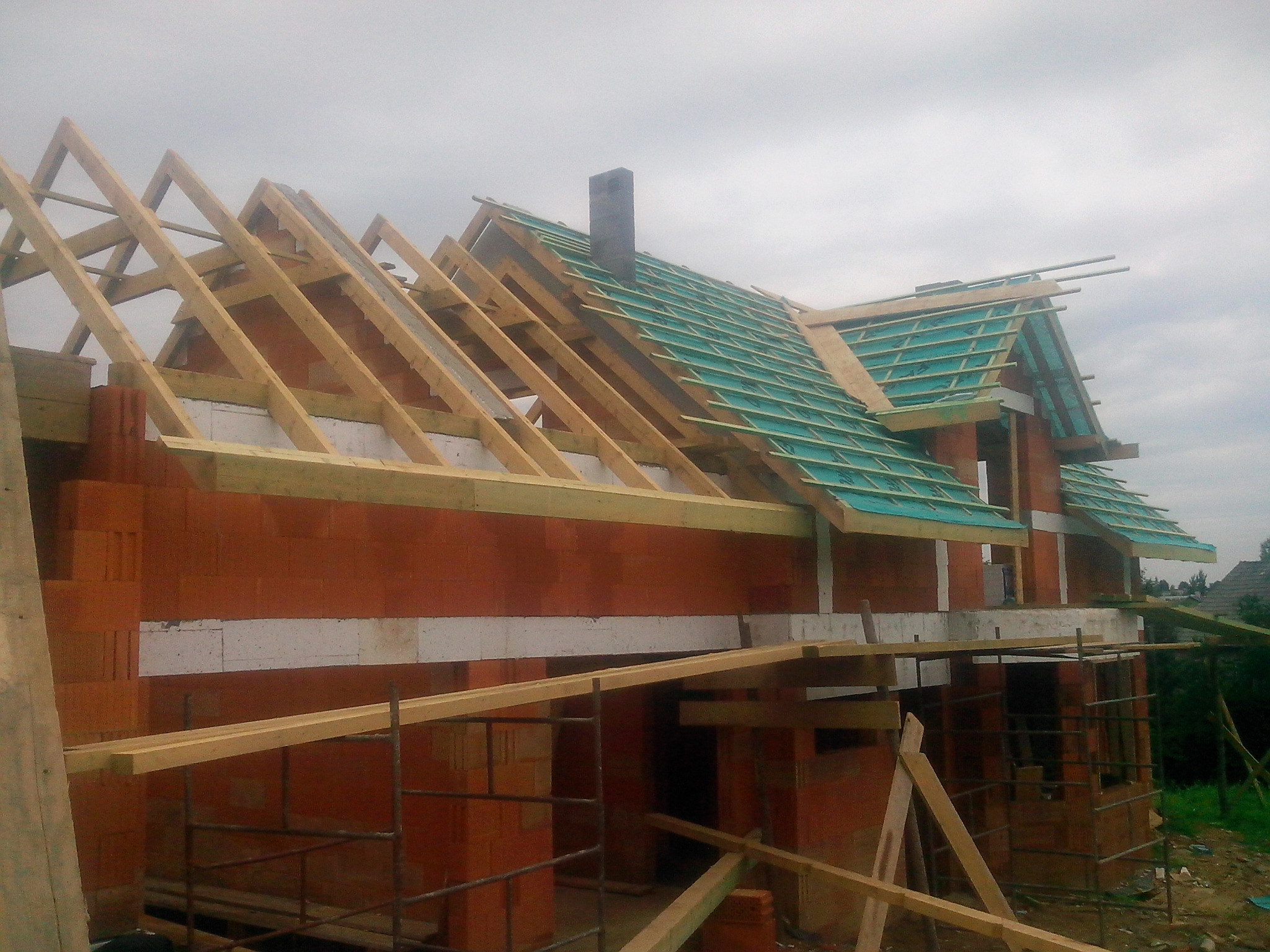 Niedokończony dom murowany z czerwonej cegły z widoczną konstrukcją dachu pokrytą folią izolacyjną, drewnianymi krokwiami i kominem, sfotografowany w pochmurny dzień.