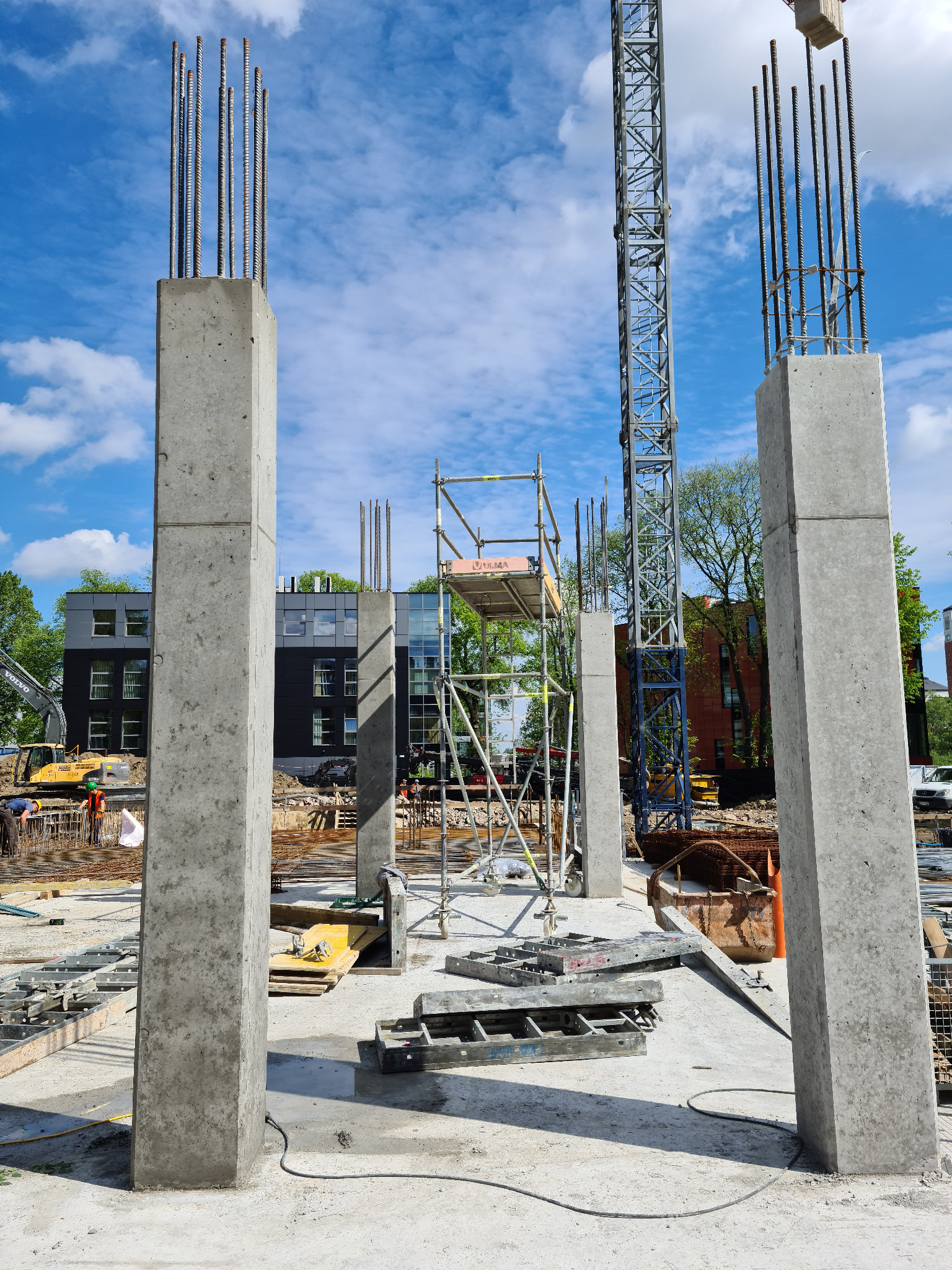 Wznoszone betonowe słupy z wystającymi prętami zbrojeniowymi na placu budowy, w tle rusztowania, żuraw budowlany i budynek mieszkalny.