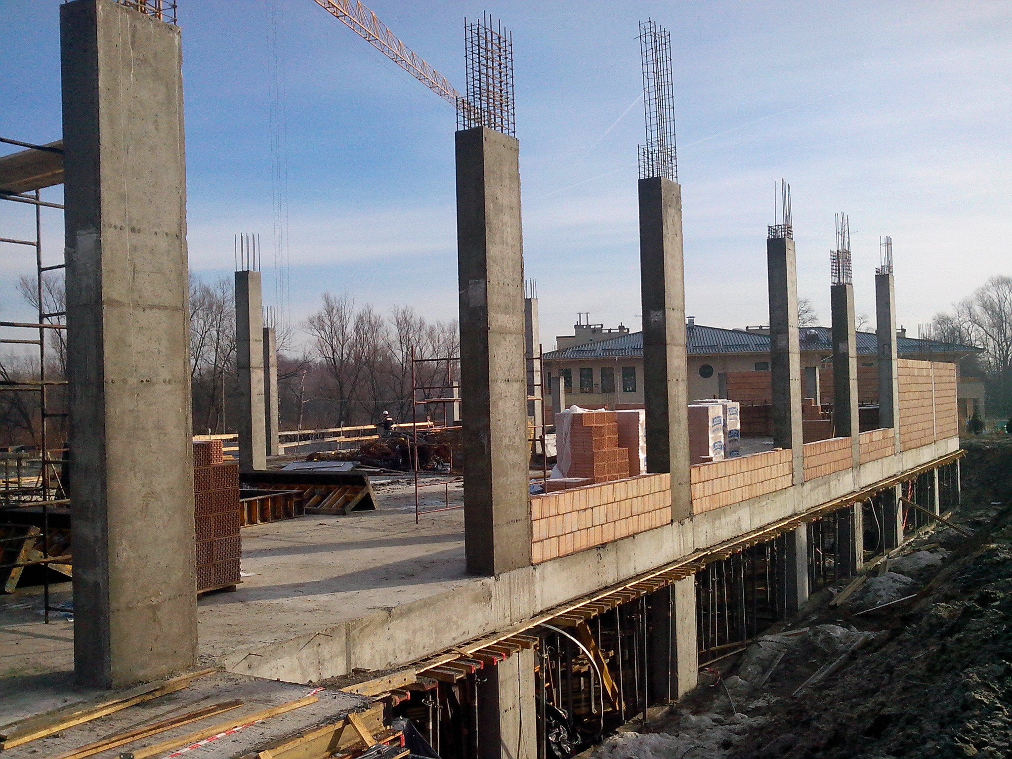Wznoszenie ścian z cegły na fundamencie domu w trakcie budowy, widoczne betonowe kolumny z wystającymi prętami zbrojeniowymi, w tle żuraw budowlany i budynek.