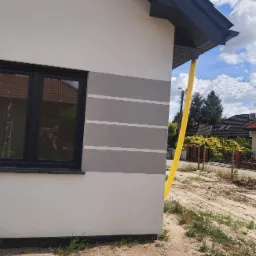 Fragment elewacji domu jednorodzinnego z widoczną aplikacją szarego tynku dekoracyjnego w pasy, czarne obramowanie okna i żółta rura spustowa przy rogu budynku, w tle widoczny ogród i domy sąsiadów.