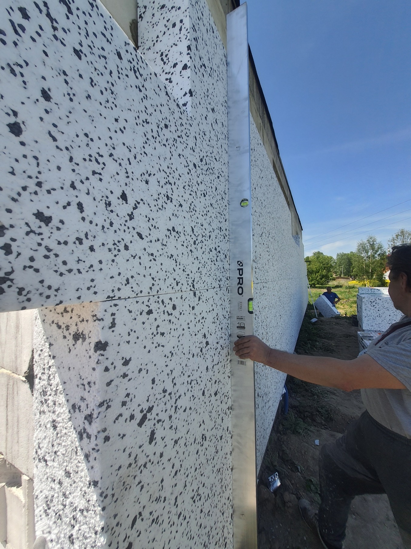 Pracownik sprawdza pionowość ocieplenia elewacji przy użyciu poziomicy PRO, widoczne płyty styropianowe w kropki.