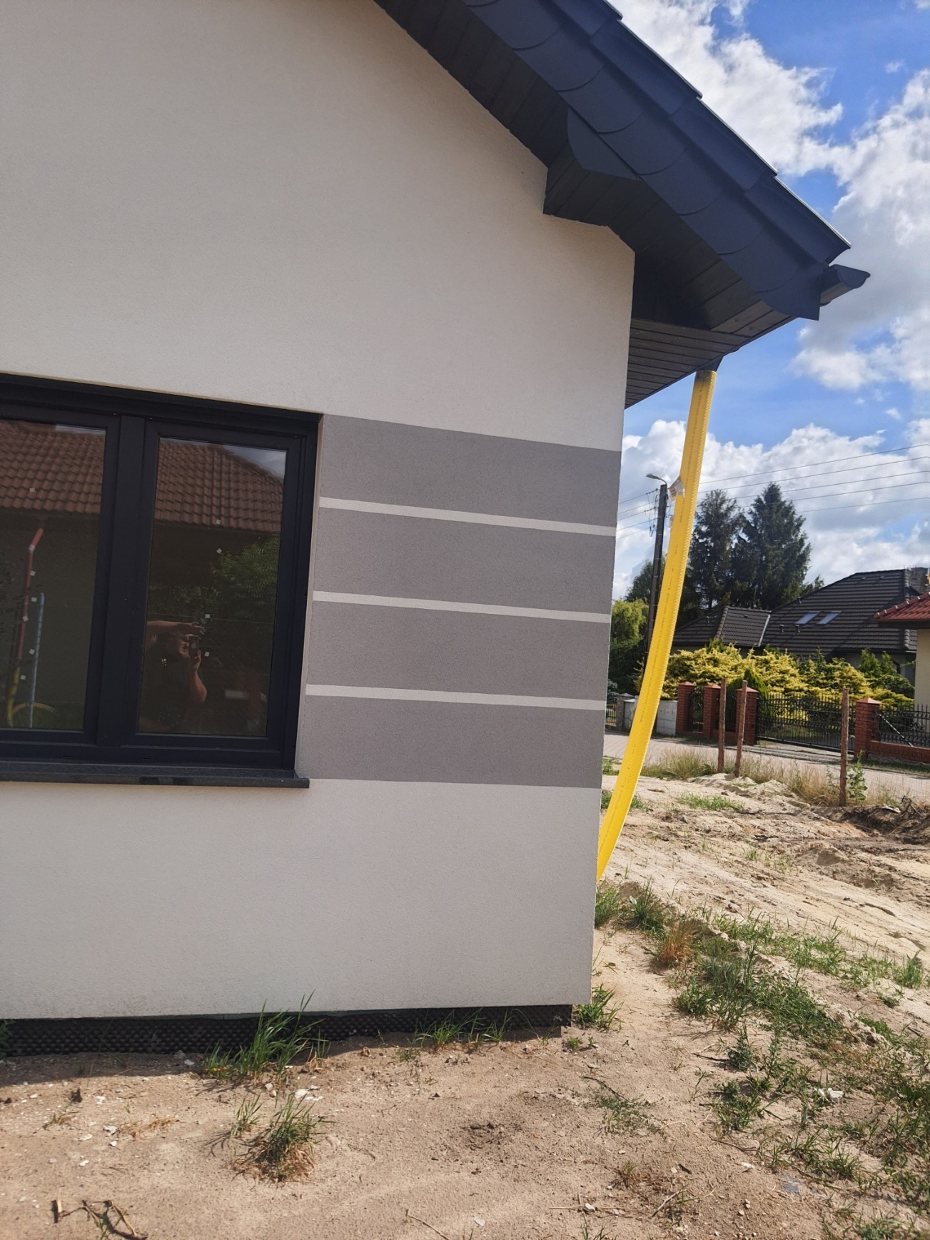 Fragment elewacji domu jednorodzinnego z widoczną aplikacją szarego tynku dekoracyjnego w pasy, czarne obramowanie okna i żółta rura spustowa przy rogu budynku, w tle widoczny ogród i domy sąsiadów.