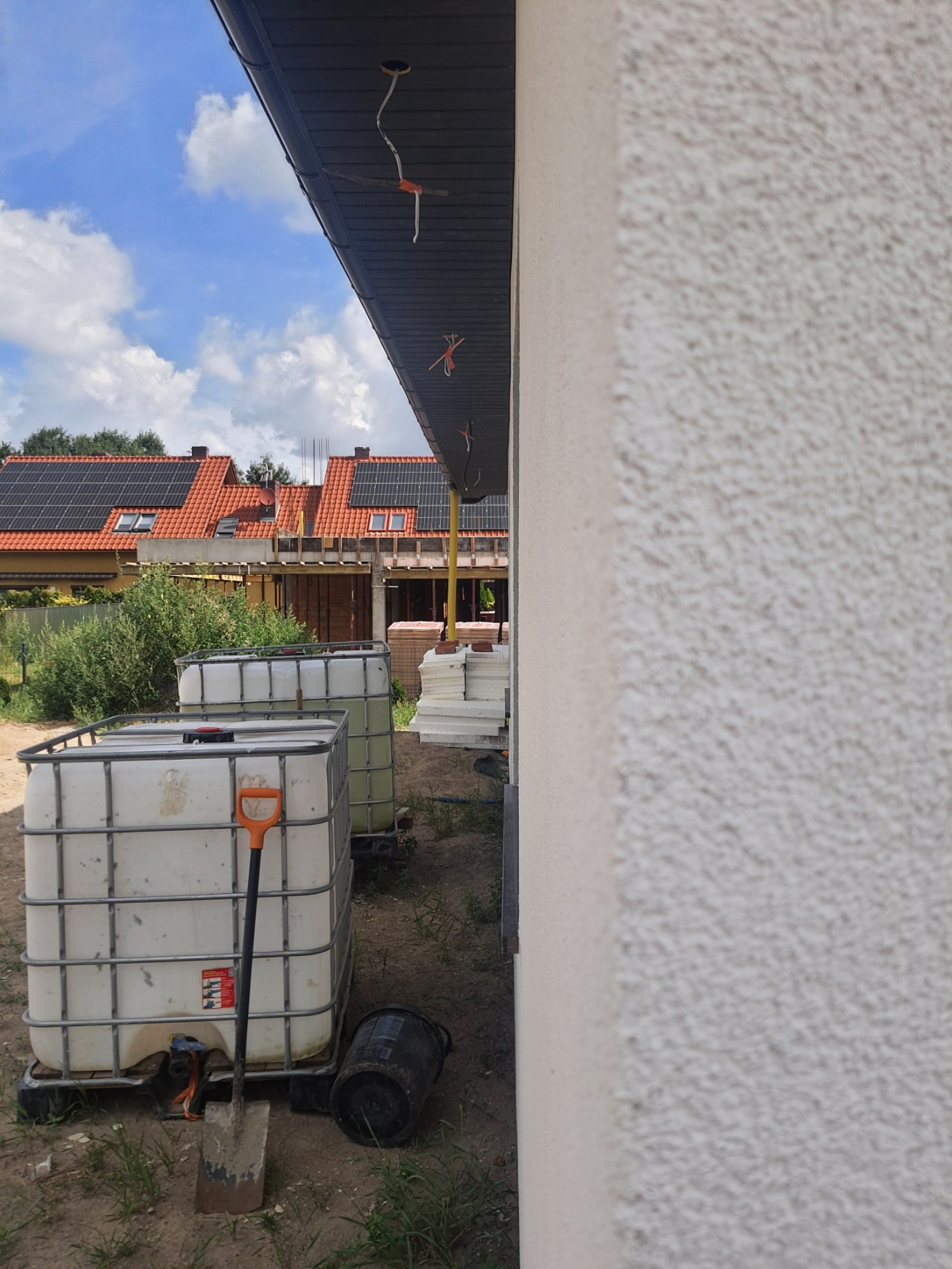 Biała, ziarnista elewacja budynku w trakcie prac, widoczne zbiorniki IBC, szpadel, wiadro, wystające kable i instalacje pod dachem, w tle domy z panelami słonecznymi.