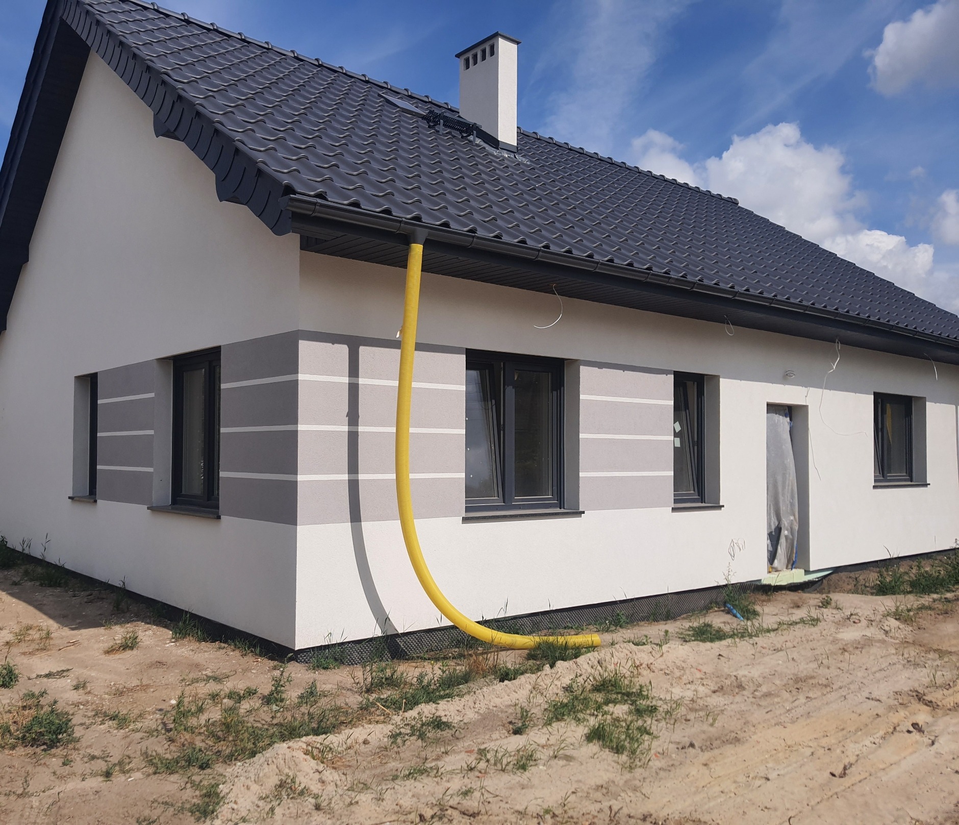 Nowoczesny dom jednorodzinny w Wałbrzychu z jasną elewacją w odcieniach szarości, ciemną dachówką i widoczną żółtą rurą wentylacyjną przy elewacji.