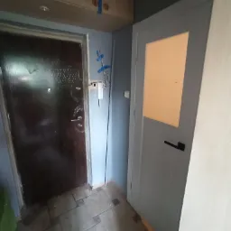 Wnętrze mieszkania z widocznymi dwoma drzwiami: ciemnymi, zewnętrznymi z widocznymi śladami użytkowania oraz wewnętrznymi, jasnymi z szybą, zamontowanymi w szarej ścianie.