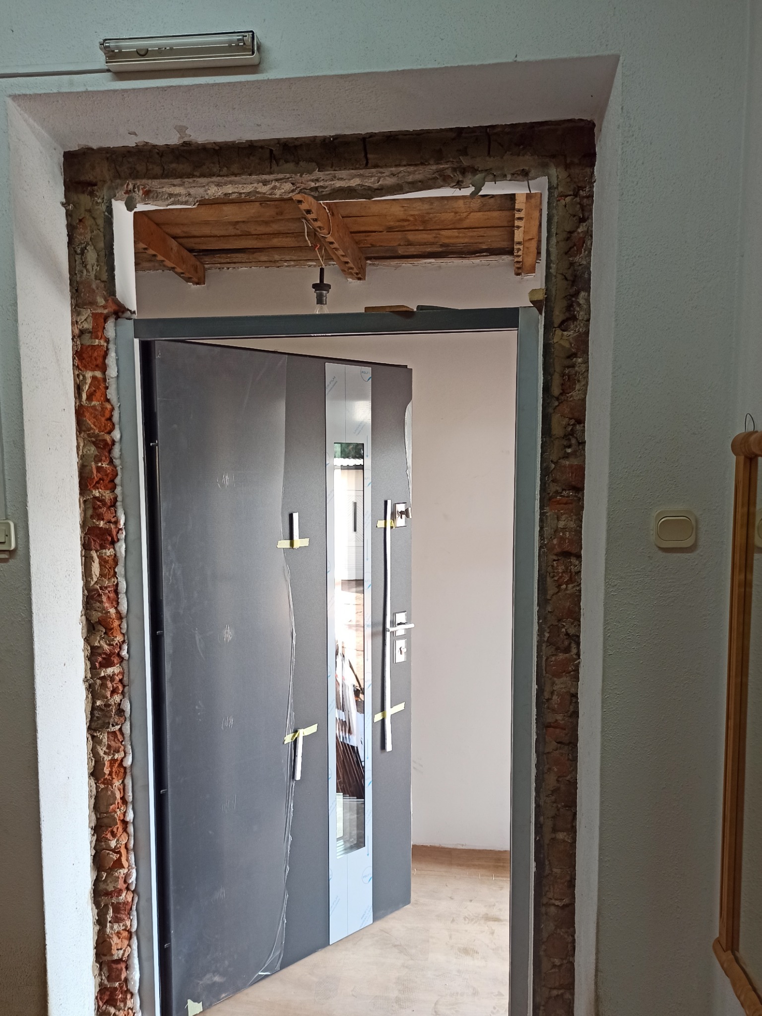 Ciemnoszare drzwi wejściowe, osadzone w surowym otworze z widoczną cegłą i pianką montażową, zabezpieczone folią ochronną, widoczne drewniane elementy nadproża.