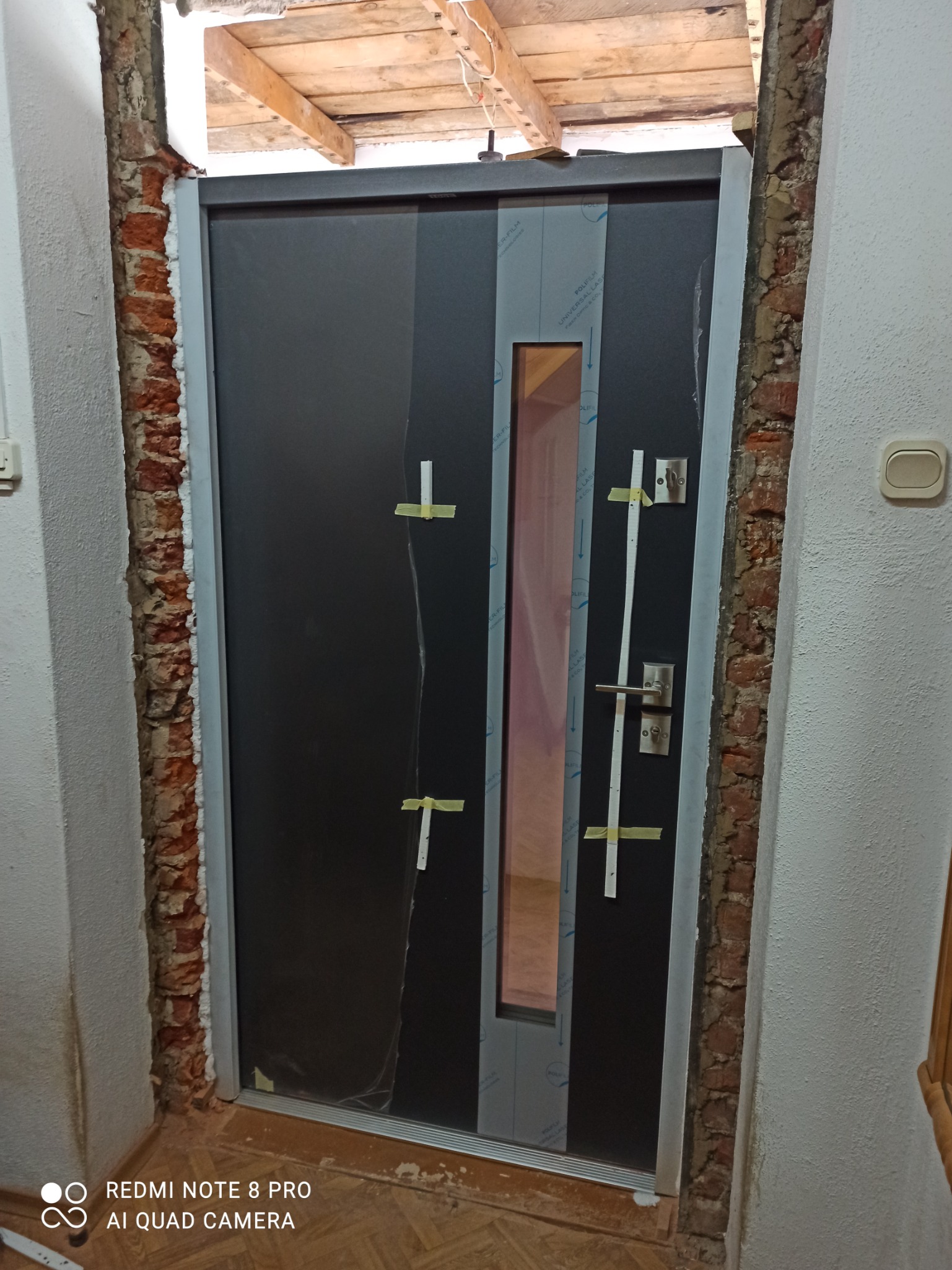 Ciemne drzwi wejściowe z szybą, zabezpieczone folią i taśmą, osadzone w surowym otworze z widoczną cegłą i pianką montażową.