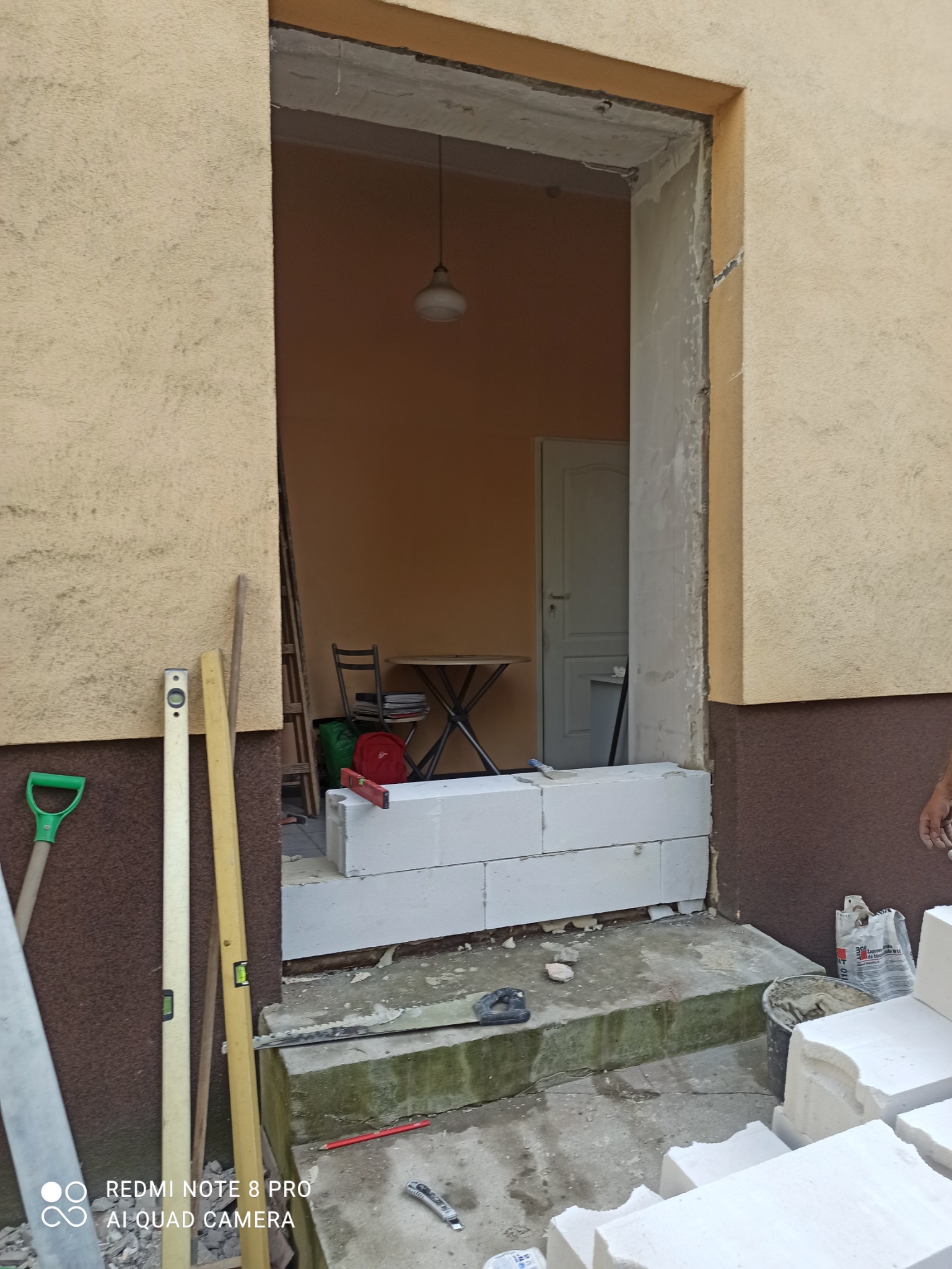 Przygotowanie otworu drzwiowego: murowanie progu z białych bloczków betonowych, widoczne narzędzia i poziomice, w tle wnętrze pomieszczenia z lampą i stołem.