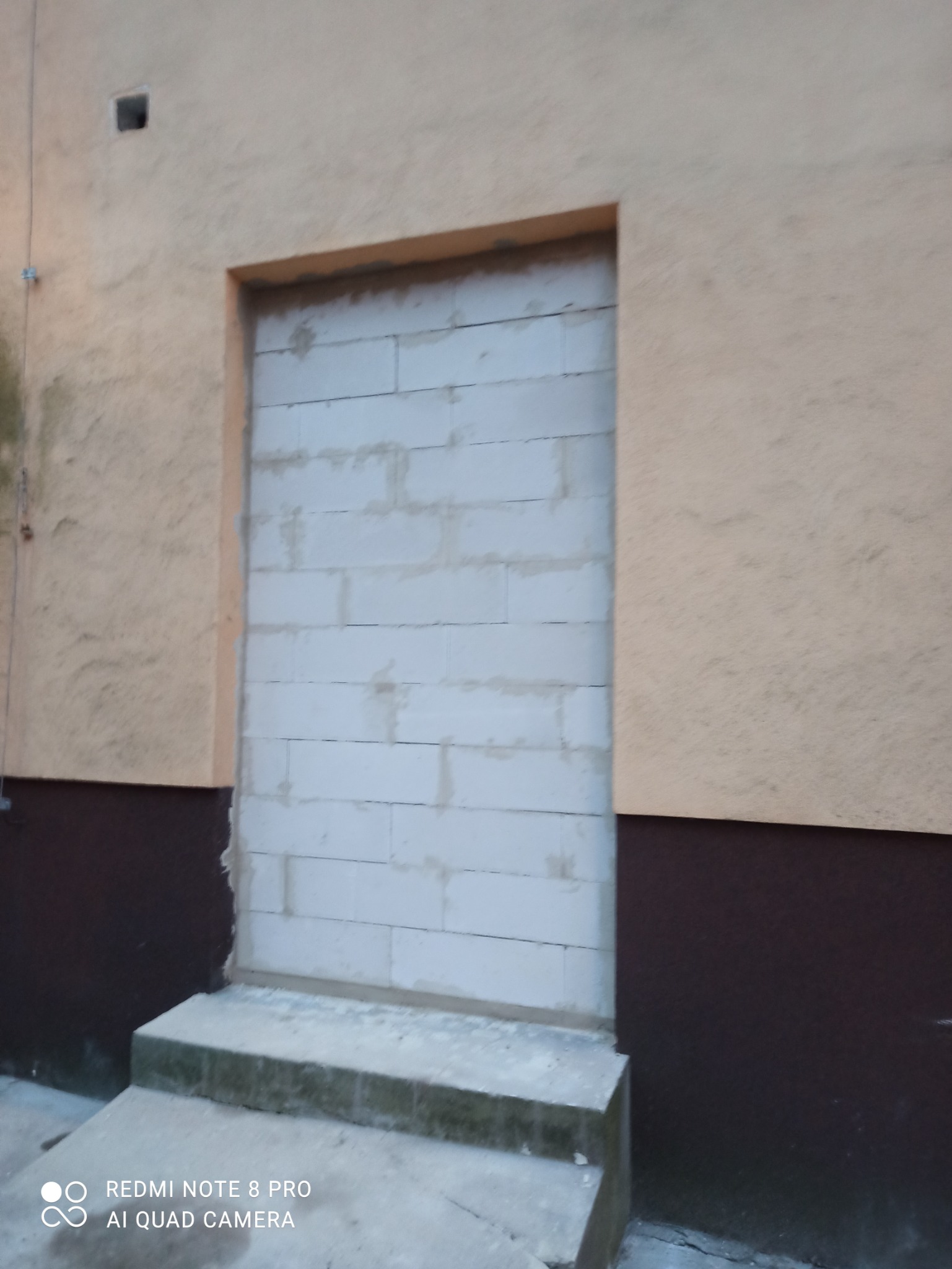 Zamknięty otwór drzwiowy w ścianie budynku, zamurowany białymi bloczkami z betonu komórkowego, widoczne schody wejściowe.