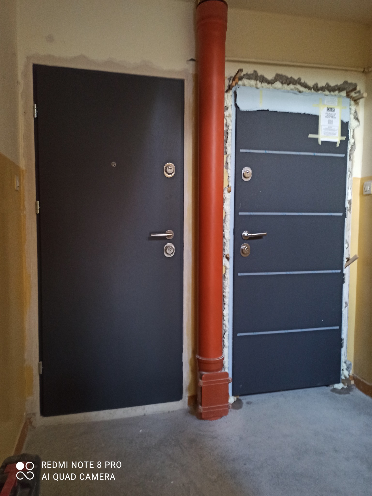 Dwa zamontowane, grafitowe drzwi wejściowe w bloku, jeden z widoczną pianką montażową i folią ochronną, obok pion kanalizacyjny.