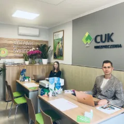 Wnętrze biura CUK Ubezpieczenia z dwoma pracownikami przy biurkach, logo firmy na ścianie, plakaty reklamowe i rośliny doniczkowe.