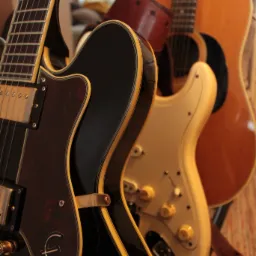 Trzy gitary: czarno-brązowa Epiphone z charakterystycznym logo, kremowa Fender Stratocaster z brązowym paskiem i akustyczna gitara w odcieniach brązu, ustawione blisko siebie w pomieszczeniu.