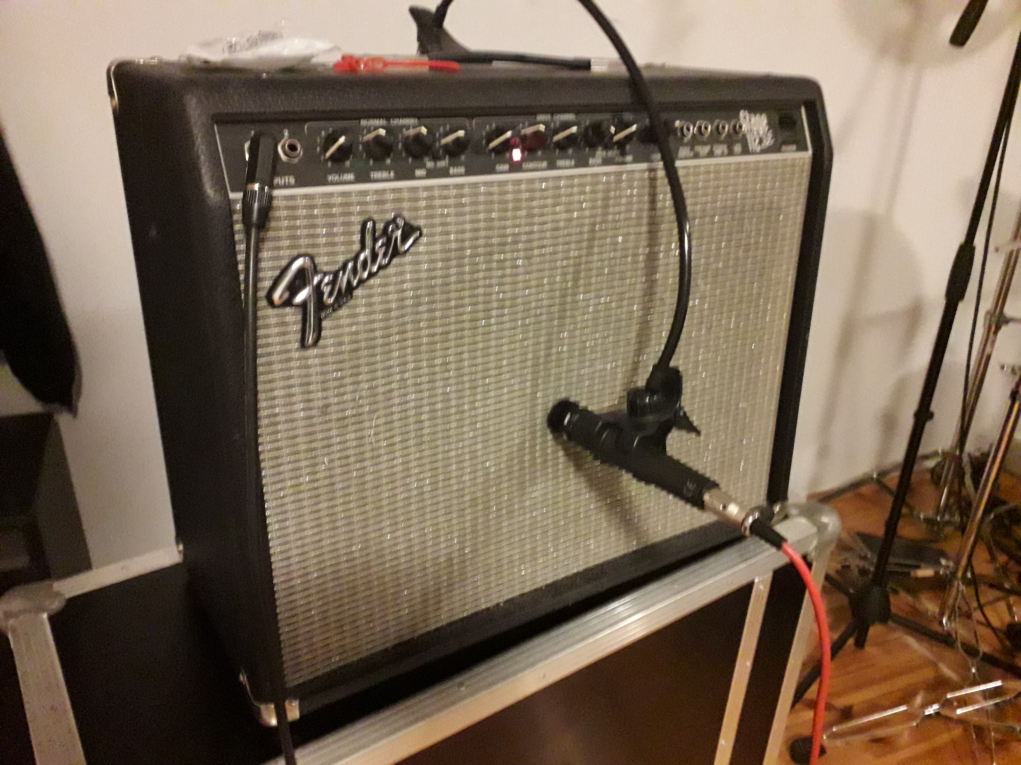 Wzmacniacz gitarowy Fender z mikrofonem przymocowanym do grilla, ustawiony na skrzyni transportowej, z widocznymi kablami i statywami w tle.