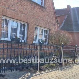 Bestbauzaune - Ciemna, metalowa konstrukcja ogrodzenia z ostrymi zakończeniami prętów, w trakcie montażu, podtrzymywana przez drewniane podpory, na tle ceglanego domu z białymi oknami i brukowanego chodnika.