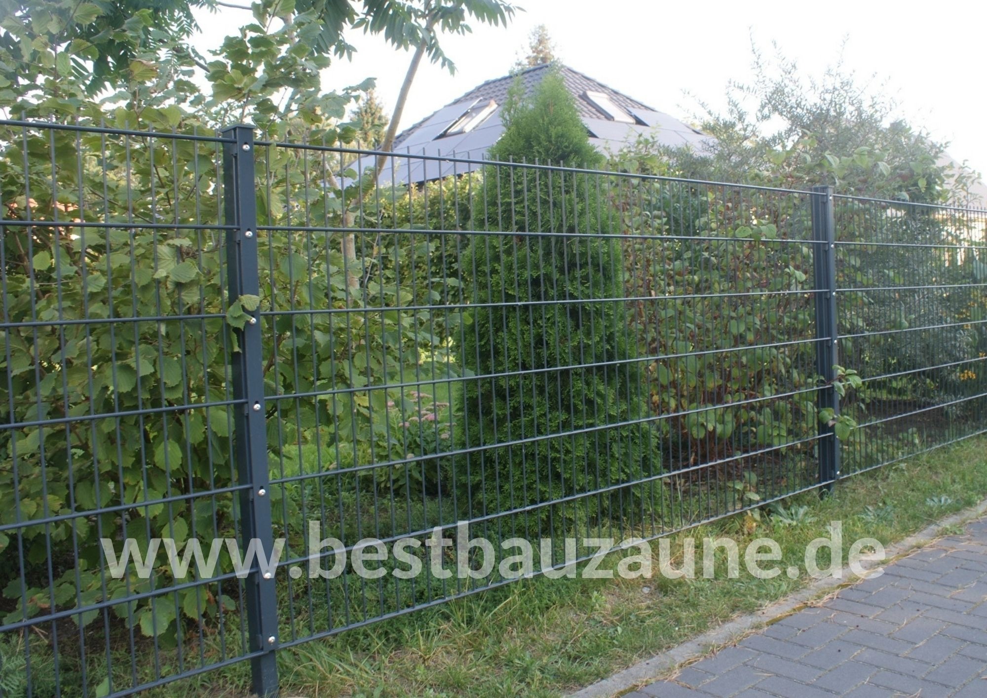 Szara, dwuprętowa konstrukcja ogrodzenia panelowego, widok z zewnątrz posesji, z zielenią w tle i fragmentem domu z panelami słonecznymi na dachu.