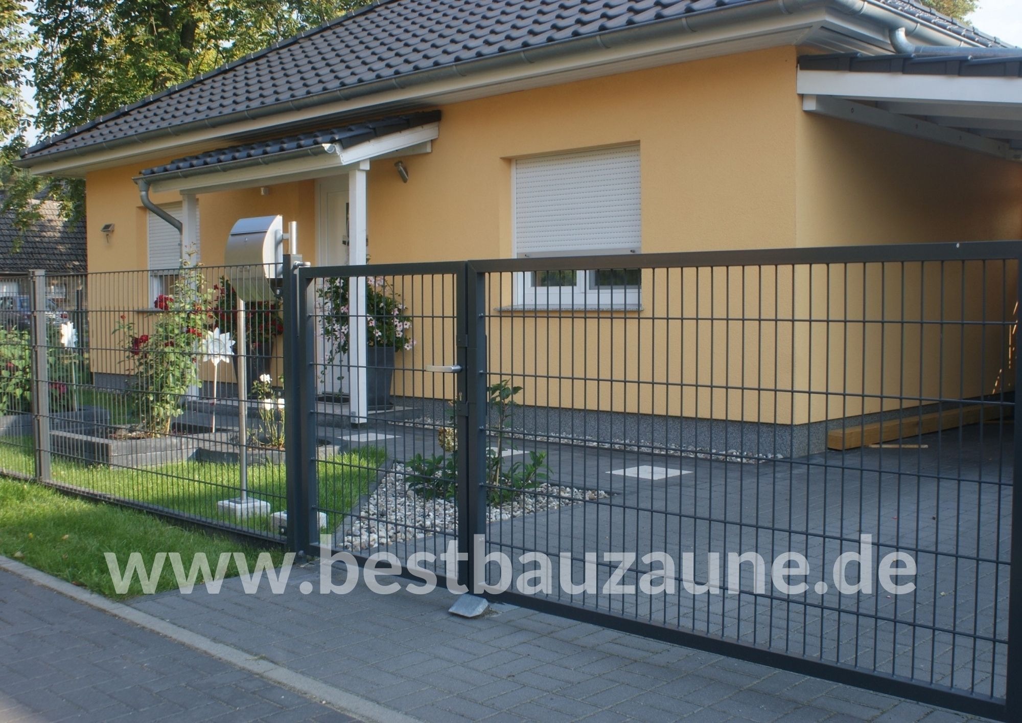 Nowoczesne ogrodzenie panelowe w kolorze grafitowym przed parterowym domem z żółtą elewacją i szarą dachówką, widoczne rośliny i trawnik.