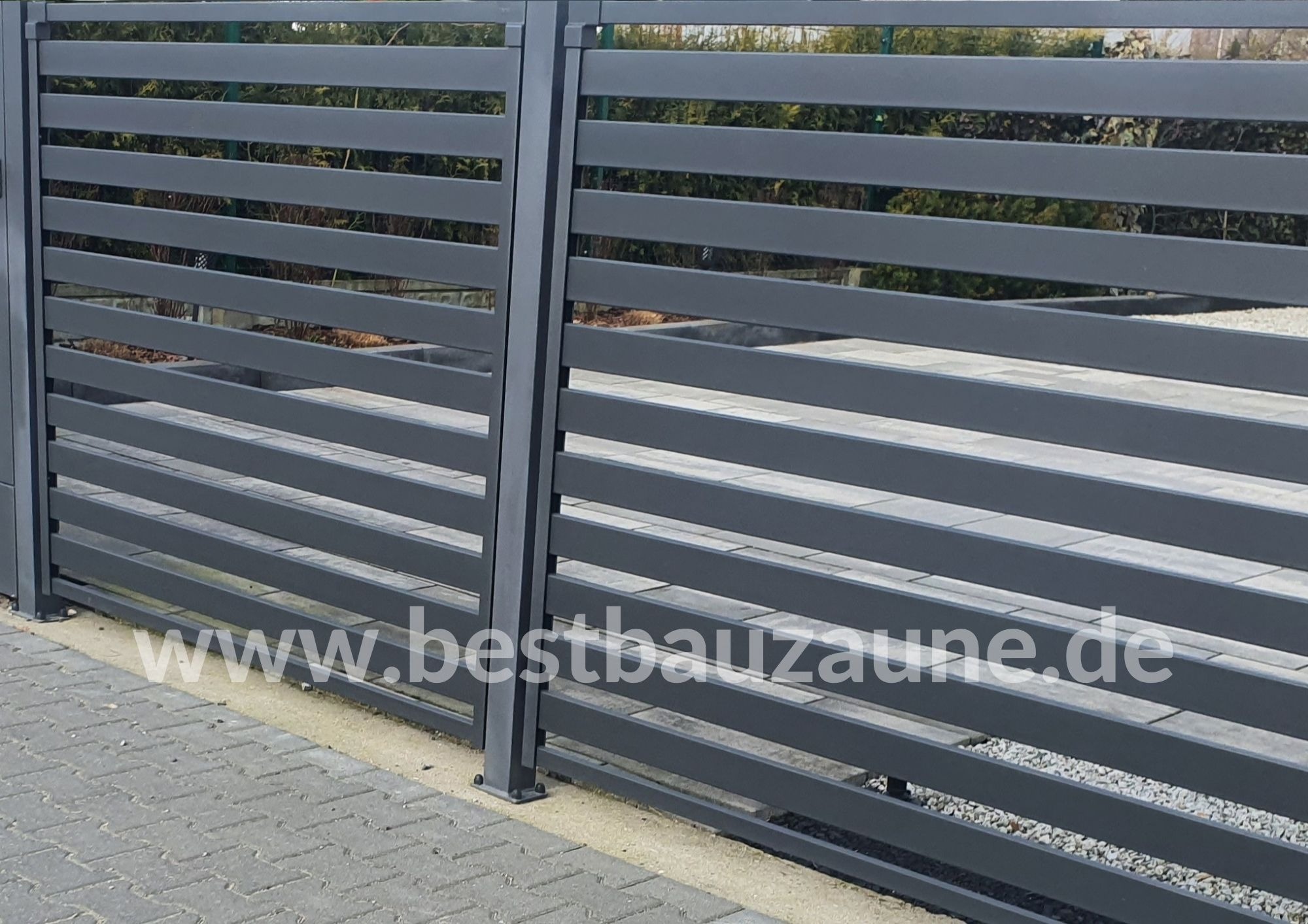 Nowoczesna, pozioma brama wjazdowa w kolorze grafitowym z widoczną kostką brukową i zielenią w tle.