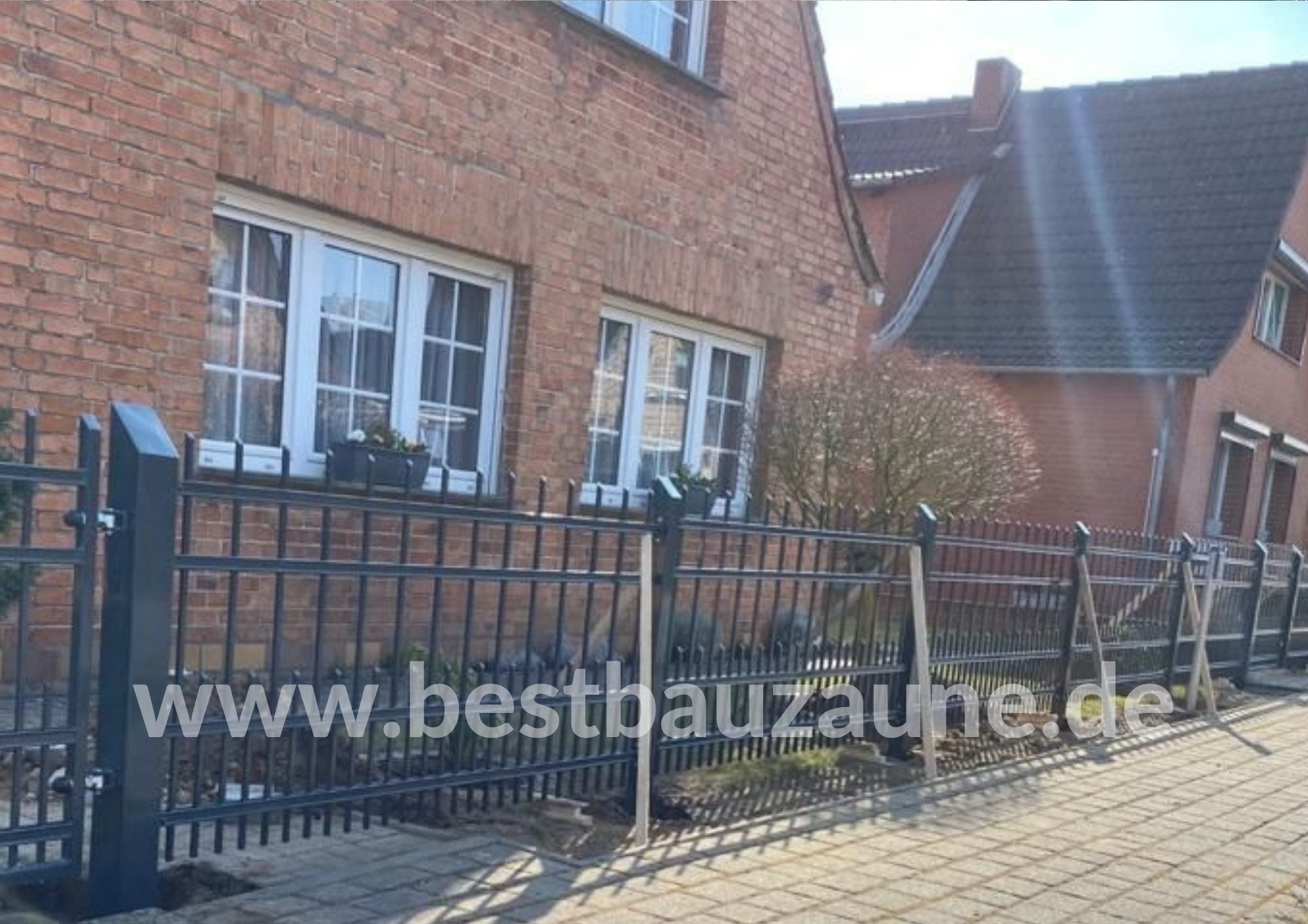 Ciemna, metalowa konstrukcja ogrodzenia z ostrymi zakończeniami prętów, w trakcie montażu, podtrzymywana przez drewniane podpory, na tle ceglanego domu z białymi oknami i brukowanego chodnika.