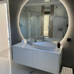 Nowoczesna łazienka z okrągłym lustrem podświetlanym LED, białą szafką pod umywalkę z ukośnymi frezowaniami, marmurową podłogą i szarą ścianą.