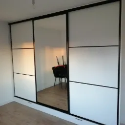 Nowoczesna szafa wnękowa z przesuwnymi drzwiami, białe fronty z czarnymi akcentami, widoczne odbicie krzesła w lustrzanym panelu, surowa żarówka zwisająca z sufitu.