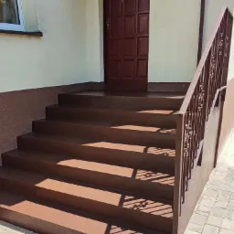 Zewnętrzne schody wejściowe pokryte brązową powłoką, z metalową balustradą, prowadzące do brązowych drzwi wejściowych.