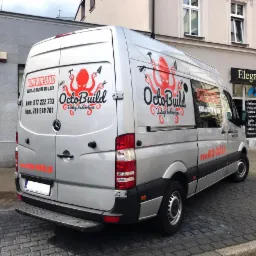 Srebrny van Mercedes z logo firmy budowlanej OctoBuild z ośmiornicą trzymającą narzędzia, zaparkowany na ulicy z kostki brukowej przed budynkiem z szyldem Eleganciki But i Sky Media.