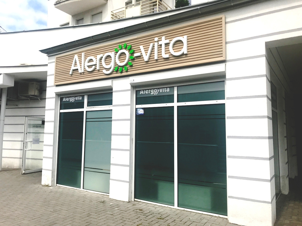 Fasada budynku z szyldem firmy Alergo-vita z logo z zielonych listków, okna z naklejkami z nazwą firmy, biała elewacja z szarymi pasami.
