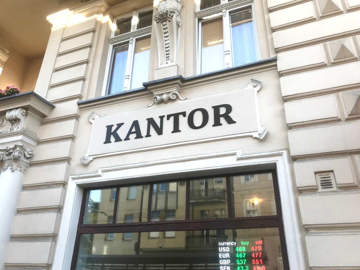 Elewacja budynku kantoru z napisem 'KANTOR' nad oknem, w którym wyświetlane są aktualne kursy walut: USD, EUR, GBP, SEK.