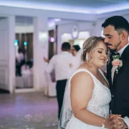 Para młoda w tańcu, panna młoda w sukni z koronkowymi aplikacjami, pan młody w ciemnym garniturze z butonierką, delikatne oświetlenie sali balowej.