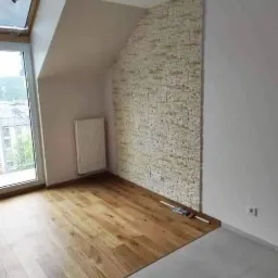 Ukończone wnętrze poddasza z drewnianą podłogą i dekoracyjną ścianą z imitacji kamienia, widoczne narzędzia pomiarowe przy ścianie, grzejnik pod oknem dachowym.