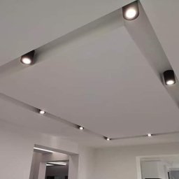 Firma Fachman - Nowoczesny sufit podwieszany z wbudowanymi, czarnymi reflektorami punktowymi, tworzącymi geometryczny wzór oświetleniowy w jasnym wnętrzu.