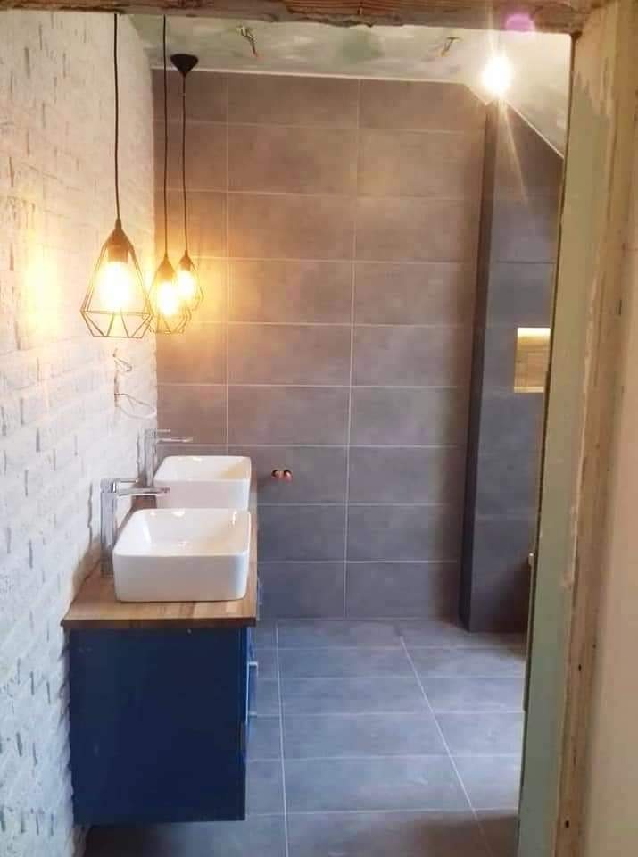 Nowoczesna łazienka z dwoma białymi umywalkami na drewnianym blacie, wiszące lampy w geometrycznych kloszach, szare płytki na ścianach i podłodze, ściana z białej cegły, widoczne niedokończone...