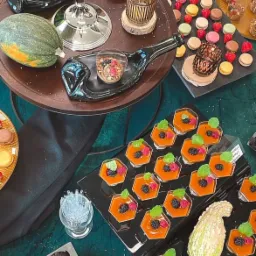 Dekoracyjny stół z różnorodnymi deserami: mini babeczki czekoladowe, musy owocowe w heksagonalnych naczyniach, makaroniki, owoce i dynie, ustawione na ciemnozielonym obrusie ze złotymi akcentami,...