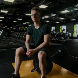 Młody mężczyzna w okularach siedzi na obciążeniu sztangi w siłowni, w tle rzędy bieżni i okna.