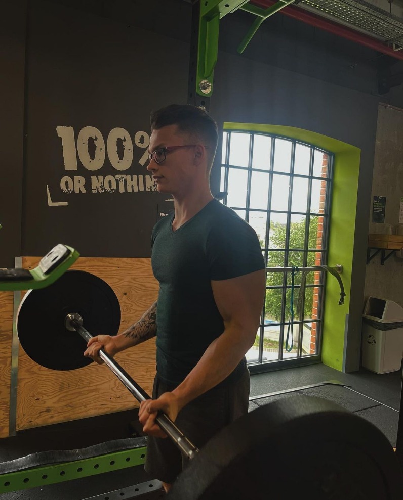 Mężczyzna w okularach podnosi sztangę na siłowni, widoczny tatuaż na ramieniu i napis '100% or nothing' na ścianie.