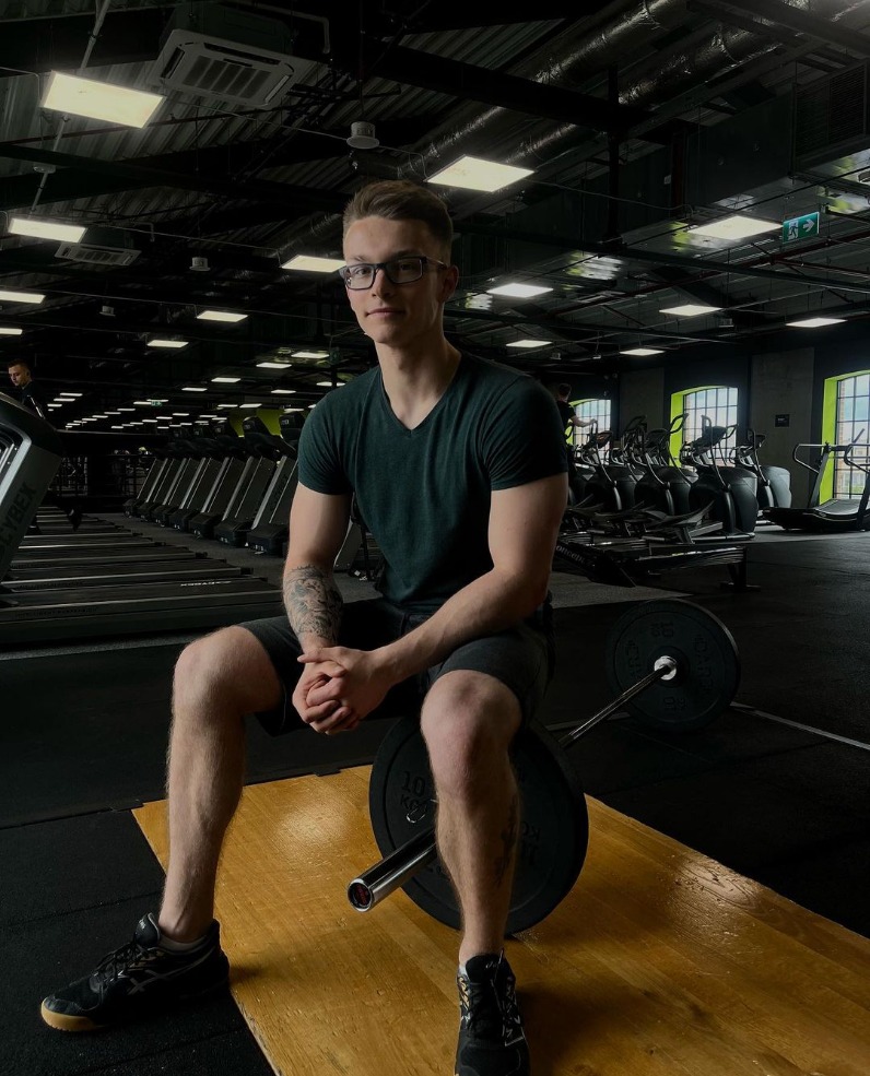Młody mężczyzna w okularach siedzi na obciążeniu sztangi w siłowni, w tle rzędy bieżni i okna.