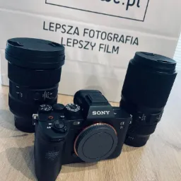 Czarny aparat Sony Alfa 7s z dwoma obiektywami na drewnianym stole, w tle torba z napisem 'Lepsza fotografia lepszy film' i logo cyfrowe.pl.