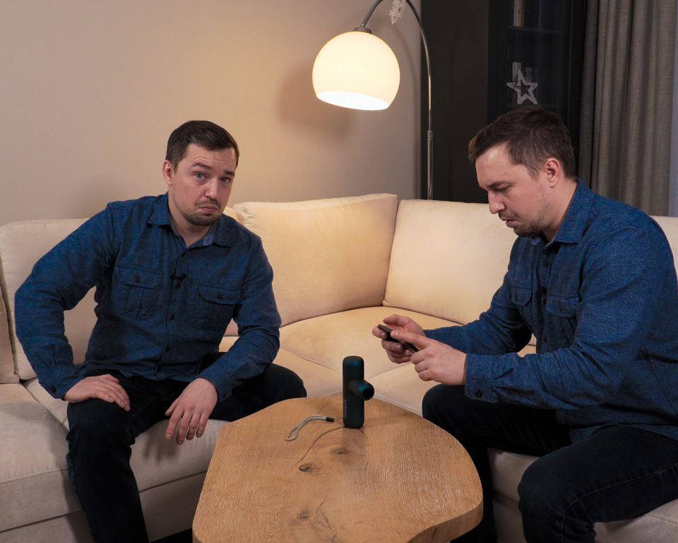 Dwaj mężczyźni siedzą na kanapie, jeden patrzy wprost z lekko niezadowoloną miną, drugi nagrywa film telefonem, na stoliku stoi masażer pistoletowy