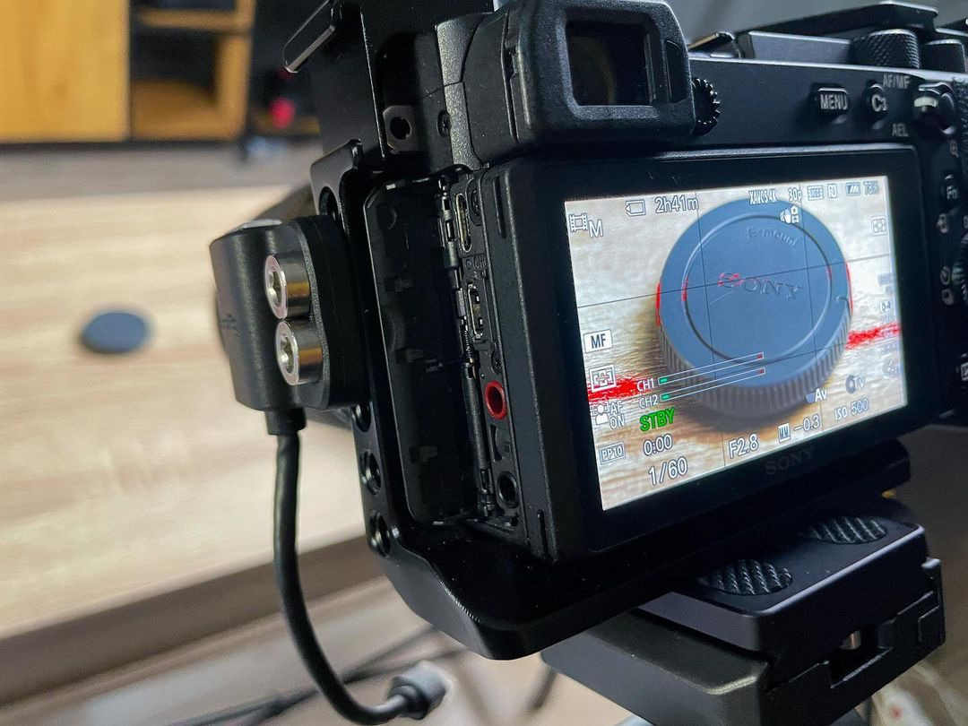 Ekran aparatu Sony z ustawieniami ekspozycji i ostrości, widoczny adapter z dwoma gniazdami audio, ustawiony na statywie.