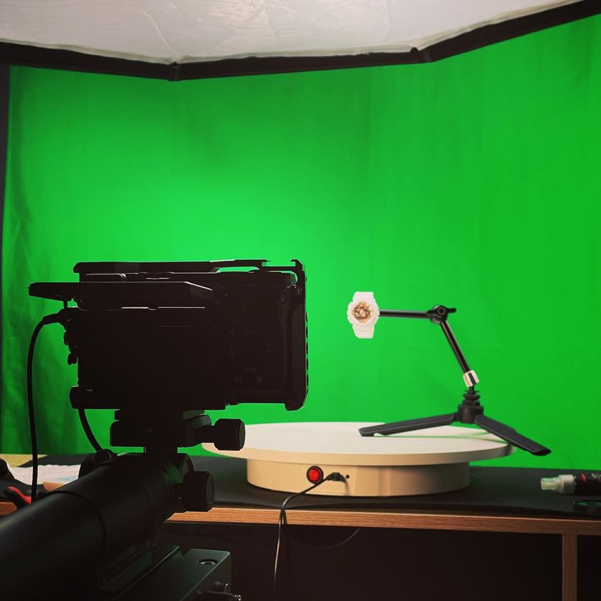 Kamera nagrywa biały zegarek na statywie, umieszczony na obrotowej platformie przed zielonym tłem w studiu filmowym.