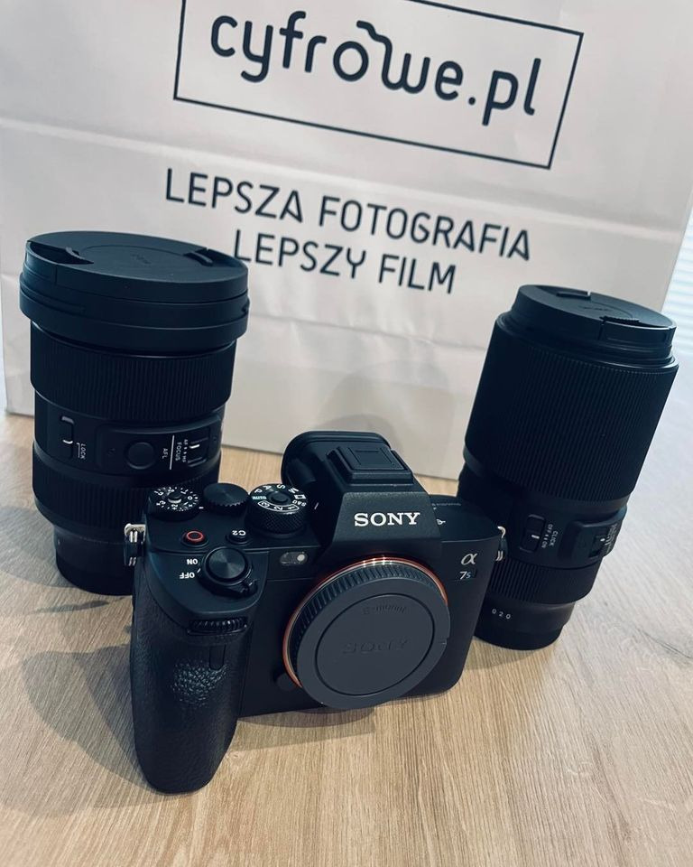 Czarny aparat Sony Alfa 7s z dwoma obiektywami na drewnianym stole, w tle torba z napisem 'Lepsza fotografia lepszy film' i logo cyfrowe.pl.