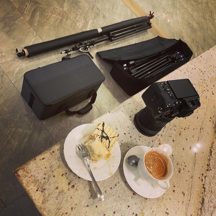Sprzęt filmowy: kamera, statywy w torbach, obok kawa i kawałek ciasta na blacie o wzorze kamienia.