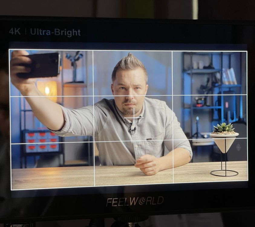 Mężczyzna z mikrofonem krawatowym, nagrywany przez monitor z siatką kompozycyjną, trzyma smartfon i robi selfie, widoczne oświetlenie studyjne i roślina w tle.