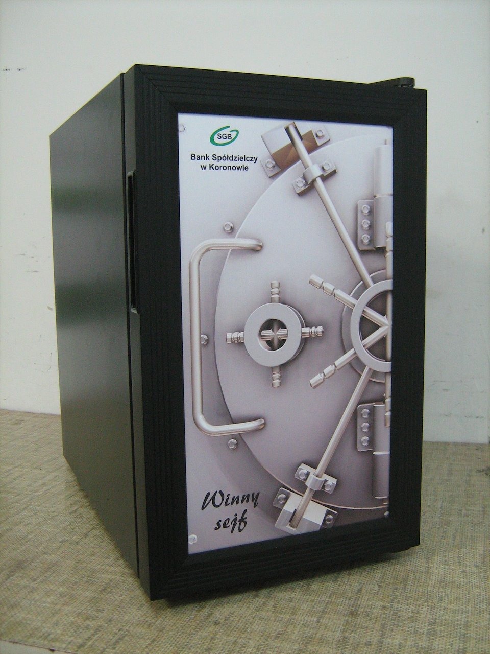 Mała czarna lodówka z grafiką sejfu bankowego i napisem 'Winny sejf' w dolnej części, logo SGB Bank Spółdzielczy w Koronowie w górnej części.