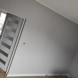 Świeżo pomalowane szare ściany w pokoju na poddaszu, widoczne gniazdka elektryczne, biała listwa przypodłogowa i nowoczesne białe drzwi z przeszkleniami.