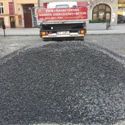 Kopuła ciemnoszarego kruszywa na placu miejskim, w tle samochód dostawczy z reklamą materiałów budowlanych i zabudowa miejska.